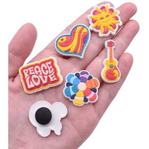 A hand holding six colourful PVC shoe charms featuring designs like a heart, sun, guitar, and flowers.