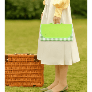 A green and checkered picnic blanket is held by a person wearing a white skirt, standing beside a wicker basket.