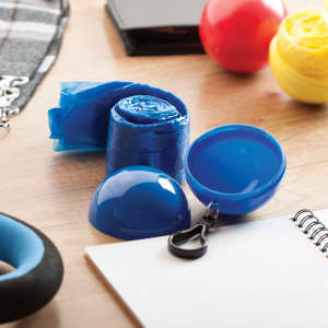 A blue keyring featuring a compact, rolled-up poncho and a snap closure, surrounded by colourful items on a table.