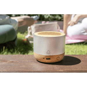 A wireless speaker with a silver top and a natural wood base, placed on a wooden surface in a grassy outdoor setting.
