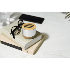 A compact white and wood wireless speaker sits on a stack of books, alongside glasses and a smartphone.