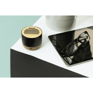 A wireless speaker in natural wood and black, positioned on a white surface next to a tablet and glasses.