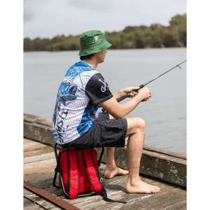 A man wearing a blue and white polyester fishing tee and a green bucket hat sits by the water, fishing while seated on a red bag.