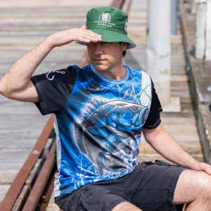 A man wearing a blue and black polyester fishing tee and a green bucket hat, sitting on a dock.