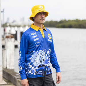 A man wearing a blue long sleeve fishing polo with a yellow collar and a matching bucket hat, beside a waterway.