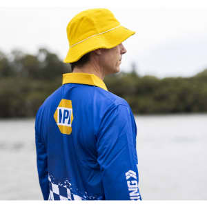 A man wearing a blue long sleeve fishing polo and a yellow bucket hat stands by the water. The shirt features a logo.