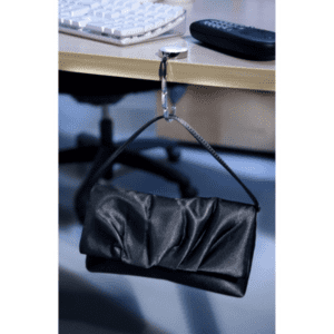 A black bag hanger clips onto a desk, supporting a black clutch. A computer keyboard and remote are in the background.