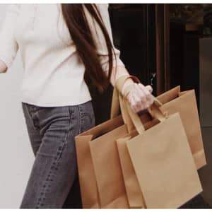 A person holding multiple brown paper bags with paper webbed handles, wearing a light-coloured top and jeans.