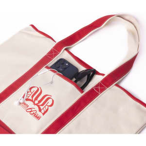 A canvas tote bag in cream and red, featuring a logo and pockets with a phone and sunglasses visible inside.