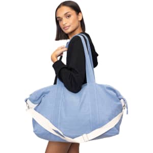 A blue duffle bag with a white shoulder strap, held by a model wearing a black hoodie. The bag has a branding logo.