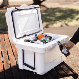 A white cooler box open with various drink bottles inside, placed on a wooden table in a park setting.