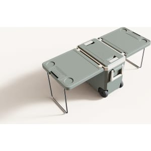 A green cooler box with folding table and chair attachments, featuring wheels and a handle for easy transport.