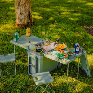 A green cooler box with a table and two chairs, surrounded by cans and snacks on a grassy area.