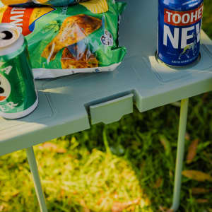 A green portable table with snacks and drinks, set on grass, featuring a can and a bottled beverage.