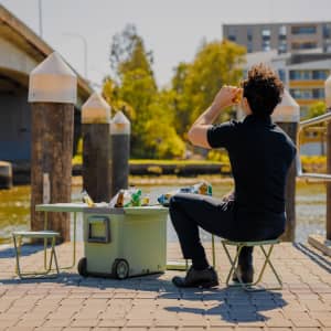A green cooler box with a table and chair, set up by a waterfront with a person sitting and enjoying a drink.