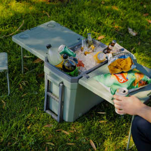 A green cooler box with a table and chair, filled with drinks and snacks, set on grass.