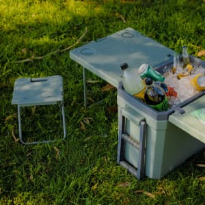 A green cooler box with a table and a small chair, filled with beverages and ice, set on grass.