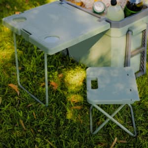 A green cooler box with a table and a small folding chair, placed on grass, featuring drink holders.