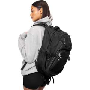 A black backpack with multiple compartments is worn on one shoulder by a person in a grey hoodie.