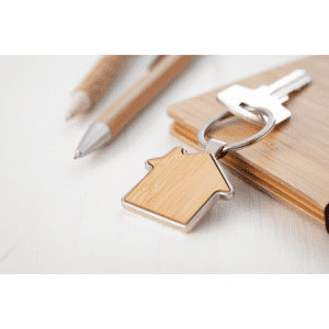 A bamboo keyring in a house shape, featuring a silver keyring attachment, placed next to pens and a wooden surface.