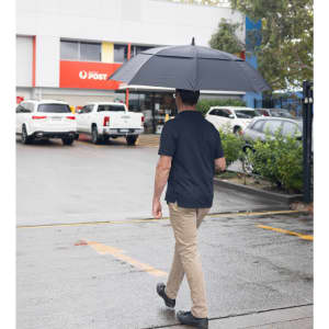 A person walking under a large black golf umbrella, with cars and a post office in the background.