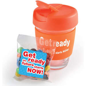 A reusable clear coffee cup with an orange lid, accompanied by a packet of M&M's.