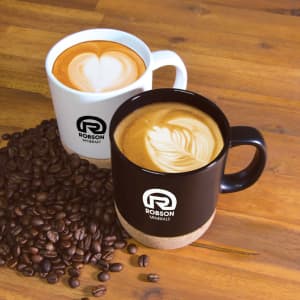 Two ceramic mugs, one white and one black with cork bases, sit beside scattered coffee beans. Both mugs feature a logo.