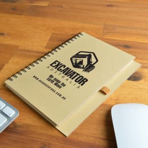 A tan spiral-bound notebook with a logo on the cover, placed on a wooden surface next to a white mouse and keyboard.