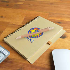 A brown spiral-bound notebook with a logo and a wooden pen resting on a wooden desk, next to a keyboard and mouse.