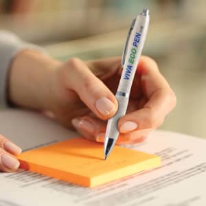 A hand holds a silver and green pen, writing on an orange sticky note with printed text below. The pen features a logo.