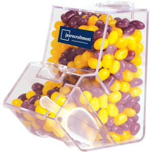 A clear dispenser filled with purple and yellow jelly beans, featuring a scoop and a branding logo.