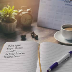 An open notebook with lined pages, a purple pen, a coffee cup, and a plant on a wooden desk.
