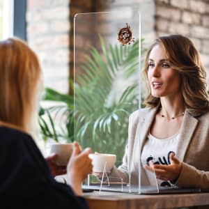 A clear acrylic personal protection screen is placed between two women seated at a table, with a plant in the background.