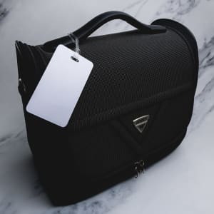 A black luggage tag with a textured surface and a handle, featuring a silver logo and a blank white tag attached.