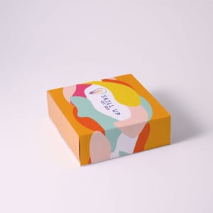 A brightly coloured gift box featuring abstract patterns and a logo on the lid. The box has a yellow background.