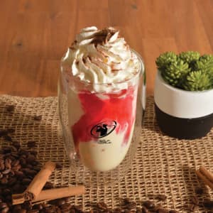 A glass cup with whipped cream, red syrup, and a base of cream, surrounded by coffee beans and cinnamon sticks.