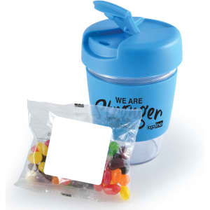 A blue reusable coffee cup with a lid next to a pack of colorful jelly beans. The cup features a logo.