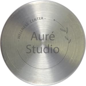 A stainless steel circular plate with a minimalist design featuring the words "Auré Studio" and a decorative leaf motif.