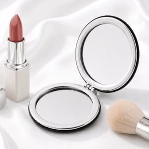 A compact mirror with a sleek silver frame, accompanied by a lipstick and a makeup brush on a soft white fabric background.