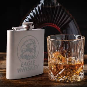 A stainless steel hip flask with an eagle design, beside a glass of whiskey with ice cubes.