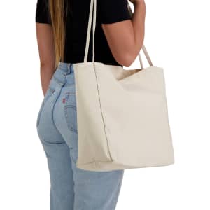 A cream-coloured tote bag is held by a person wearing a black top and blue jeans. The bag has a simple design and a logo.