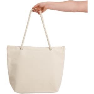 A hand holds a large, cream-coloured canvas tote bag with rope handles. The bag is plain and unbranded.