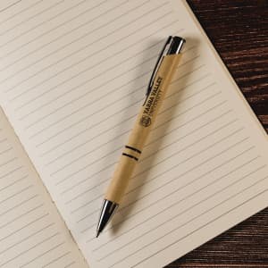A brown paper pen with a black clip and tip rests on a lined notebook page. The pen features a logo.