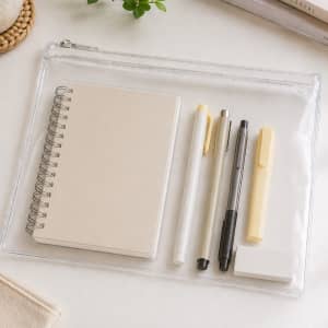 A clear pencil case containing a notebook, pens, a highlighter, and an eraser, all in neutral colours.