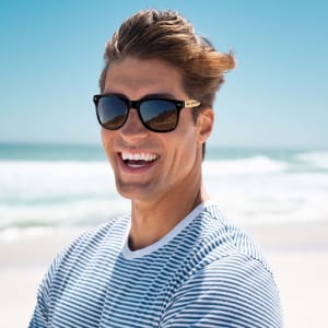 Bamboo sunglasses with dark lenses, worn by a smiling man at the beach against a bright blue sky and ocean backdrop.