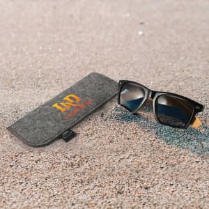 A grey case with a logo and a pair of black sunglasses resting on sandy ground.