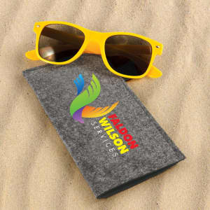 A pair of yellow sunglasses rests on a grey case, set against a sandy background. The case features a logo.