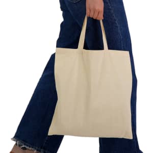 A cream-coloured tote bag with short handles, held by a person wearing blue denim jeans. The bag has a logo.