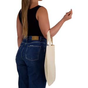 A person carrying a natural-coloured tote bag with short handles while wearing a black top and denim jeans.