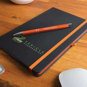 A black notebook with an orange band and an orange pen on a wooden surface, featuring a logo.
